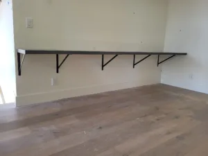 Black floating shelf with metal bracket supports mounted above baseboard on an interior wall.
