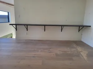 Long black floating shelf with bracket supports mounted on a wall near a window in a vaulted room.
