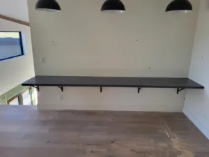 Long black floating shelf with visible brackets, mounted below three pendant lights in a vaulted room.