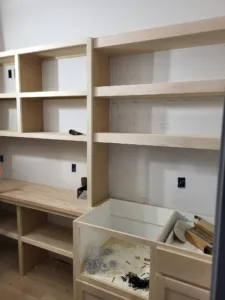 Built-in bookcase under construction with open shelving above and lower cabinet storage, raw wood frame.