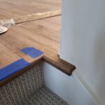 Stained wood baseboard installed at wall-to-carpet transition with protective tape on carpet.