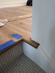 Stained wood baseboard at a wall-to-carpet transition, protective tape on the floor nearby