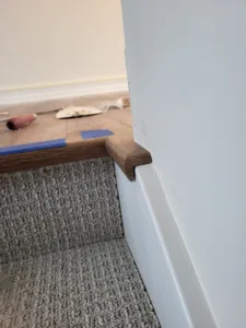 Rounded wood cap trim installed at a wall corner above carpeted stairs, blue tape marking an edge nearby.