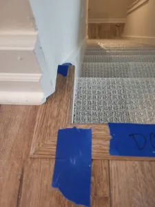Floor transition trim mitered at a corner where wood-look flooring meets carpeted stairs, tape in place.