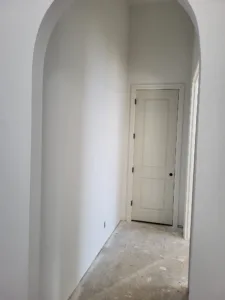 Paneled white door visible down a hallway framed by an arched opening in the foreground, unfinished floor.