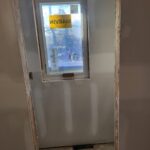 Window unit installed in rough opening with protective tape; framing and drywall visible around opening.