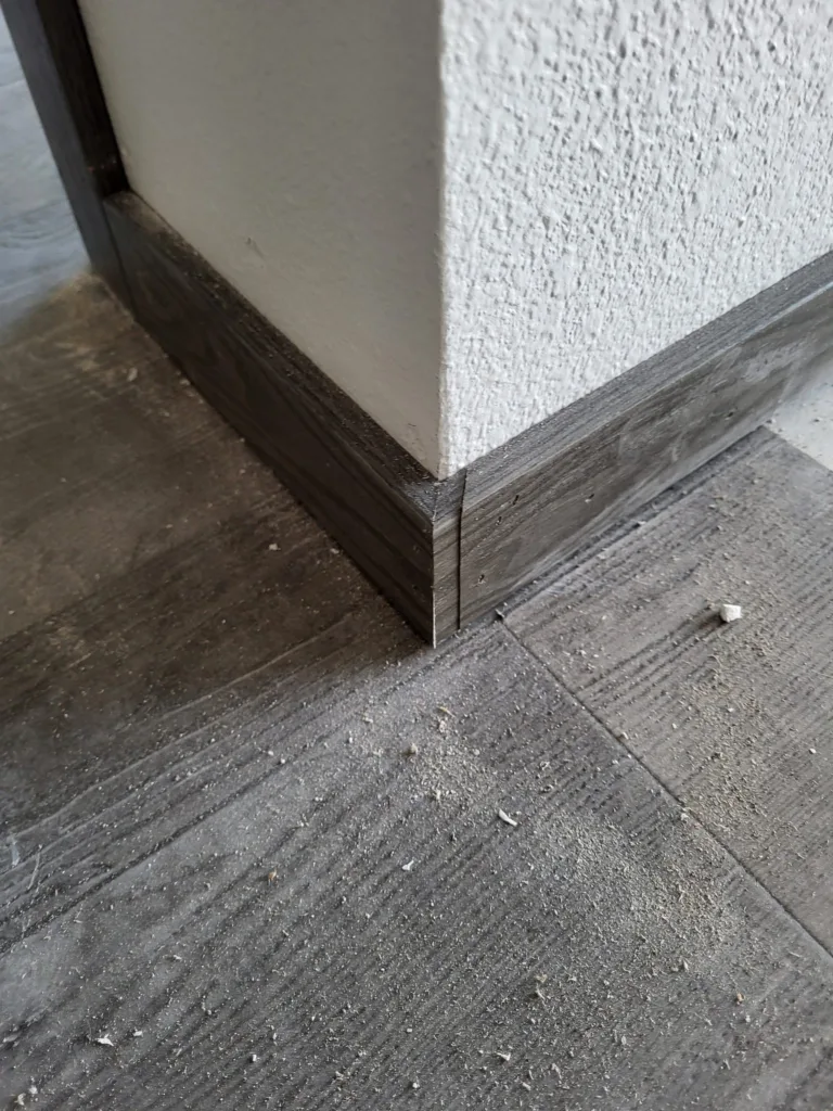 Dark stained baseboard joined at an outside corner above gray plank flooring.