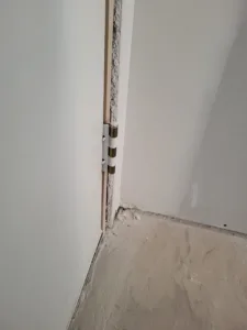 Door jamb corner with a hinge pin meeting an unfinished floor, drywall stage