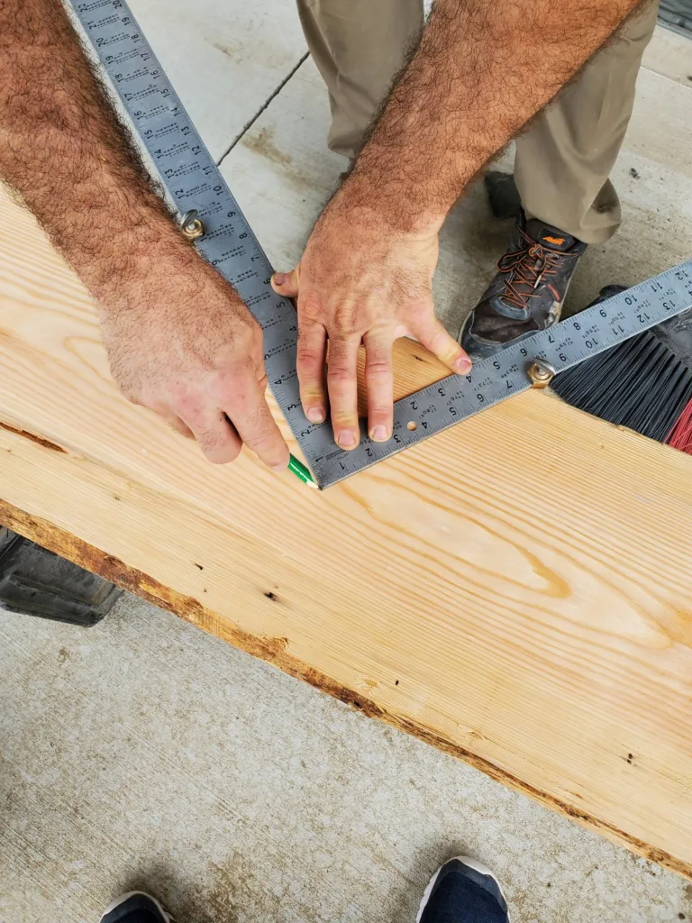 Framing square with stair gauges held on a 2x12 while a riser and tread line are traced in pencil